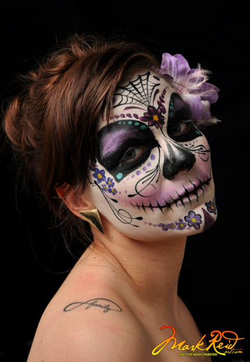 brunette woman in a highly stylized full face painting of a scull with purple highlights and the model has a matching flower behind her ear